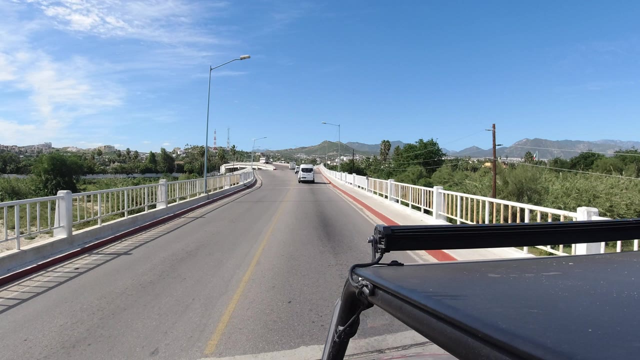 Forward car-mounted view in Los Cabos with the road bending slightly left, a white vehicle ahead, bridge railings on both sides, hills beyond, and no visible people.