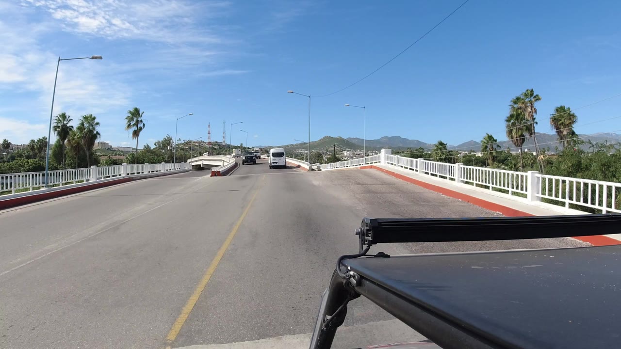 One second into the Los Cabos drive, the camera remains centered on the bridge lane with a white van ahead, palms to the right, hills on the horizon, and no visible people.