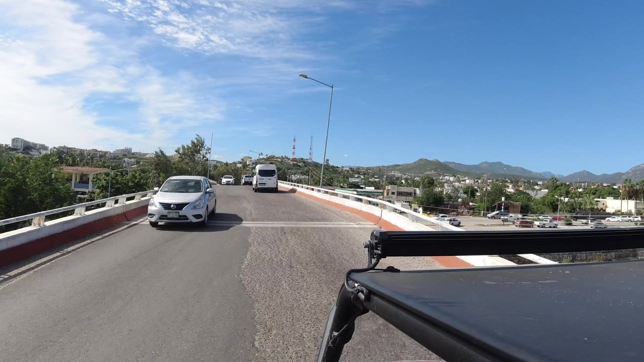 A silver car drives toward the camera on a raised Los Cabos road with a white van ahead, hillside buildings in the distance, a dark vehicle edge in the foreground, and no visible people.