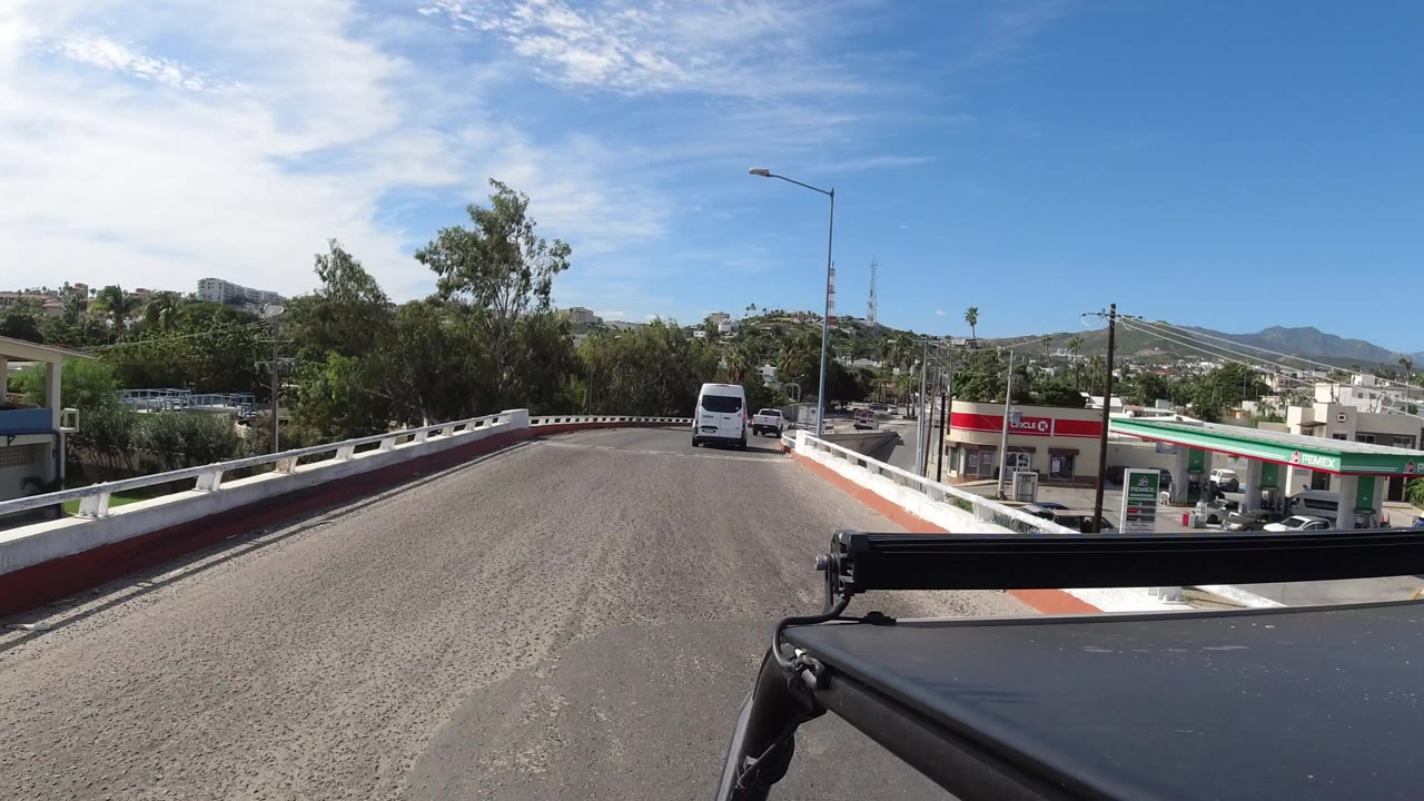 Opening frame of a Los Cabos road view from a moving vehicle, with a white van ahead, a gas station on the right, green trees, distant hills, blue sky, and no visible people.