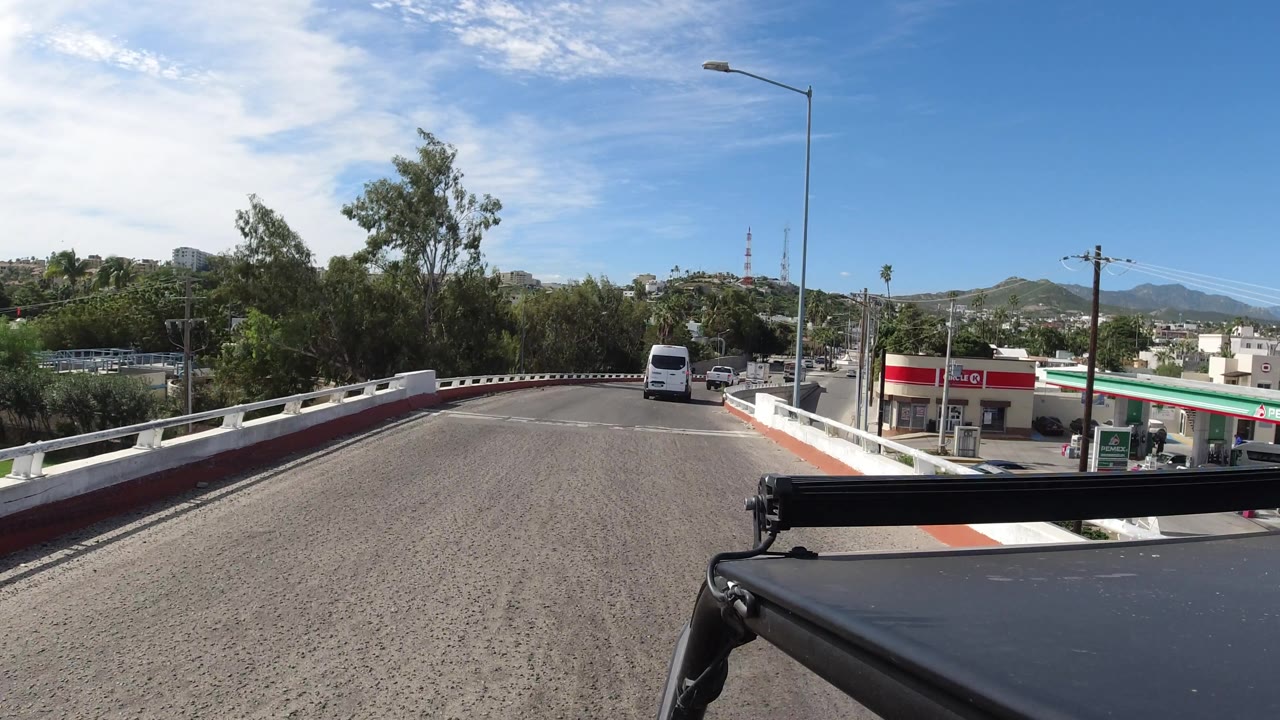 Curving road with white van ahead, utility poles, gas station, and hillside background.