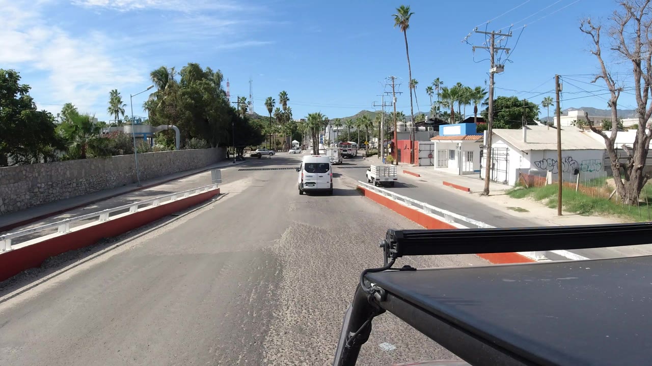 One second into the clip, the Los Cabos road remains centered with the white van slightly farther ahead, the stone wall continuing left, white and red buildings to the right, and no clearly visible people.
