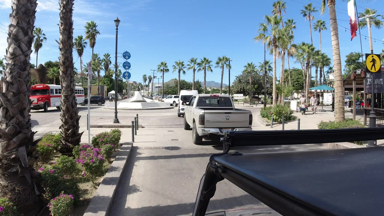 The camera remains behind a white pickup in Los Cabos, Mexico, with the black foreground vehicle edge, palm trunks, a white roundabout monument, and traffic signs visible.