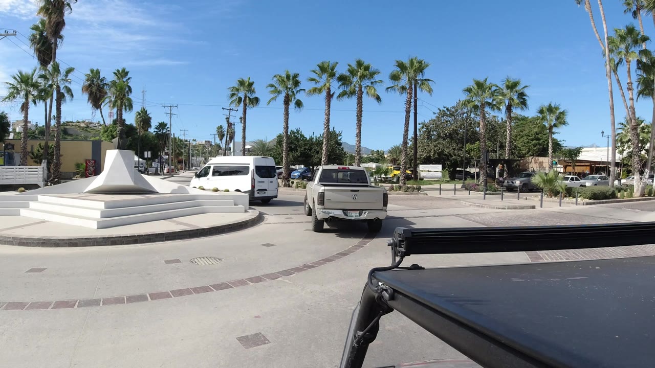 Los Cabos road scene one second in, with the camera viewpoint behind a vehicle roof, a white pickup centered in traffic, a white van to the left, palms, buildings, and distant pedestrians.