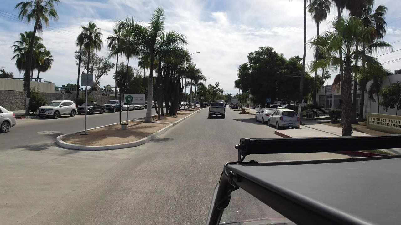 The jeep-mounted camera continues along a Los Cabos roadway past palm trees, parked vehicles, a roadside sign, and low commercial buildings, with no visible people.