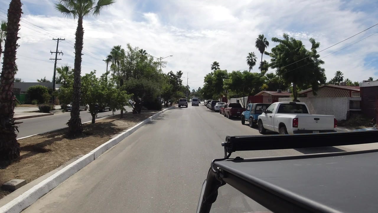 Los Cabos roadway seen from a moving vehicle, with a white pickup on the right, palms on both sides, low buildings, utility wires, and no clearly visible people.