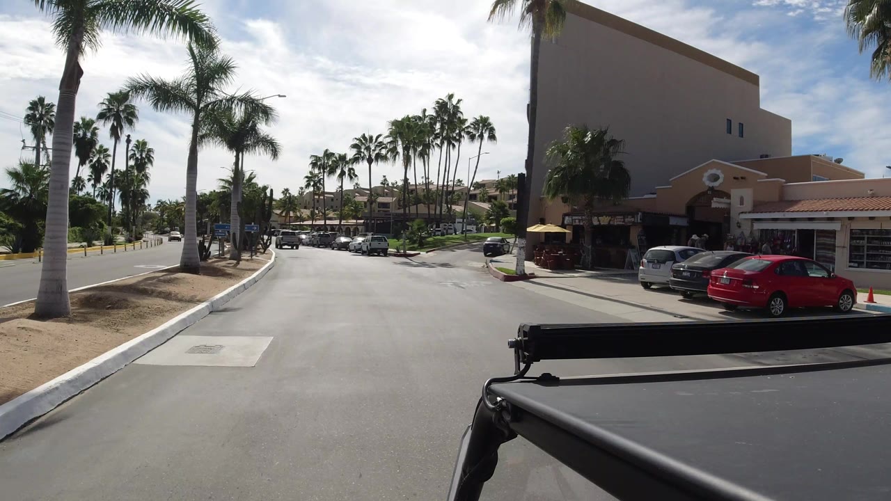 Los Cabos roadway seen over dark vehicle hardware, with a large stucco building on the right, parked cars, palms, sidewalk, and no clearly visible people.