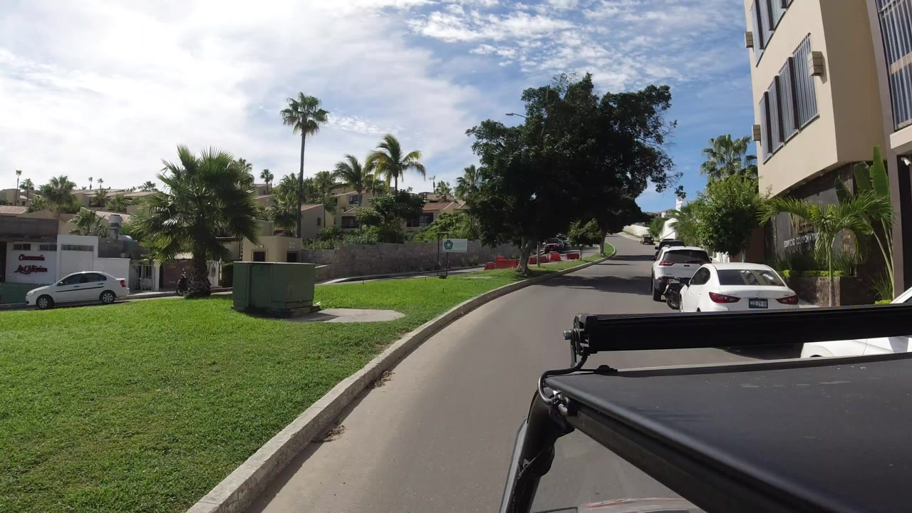 Street-level driving view in Los Cabos, Mexico, showing a paved residential road, green grass, palm trees, parked cars, and no visible people at the opening moment.