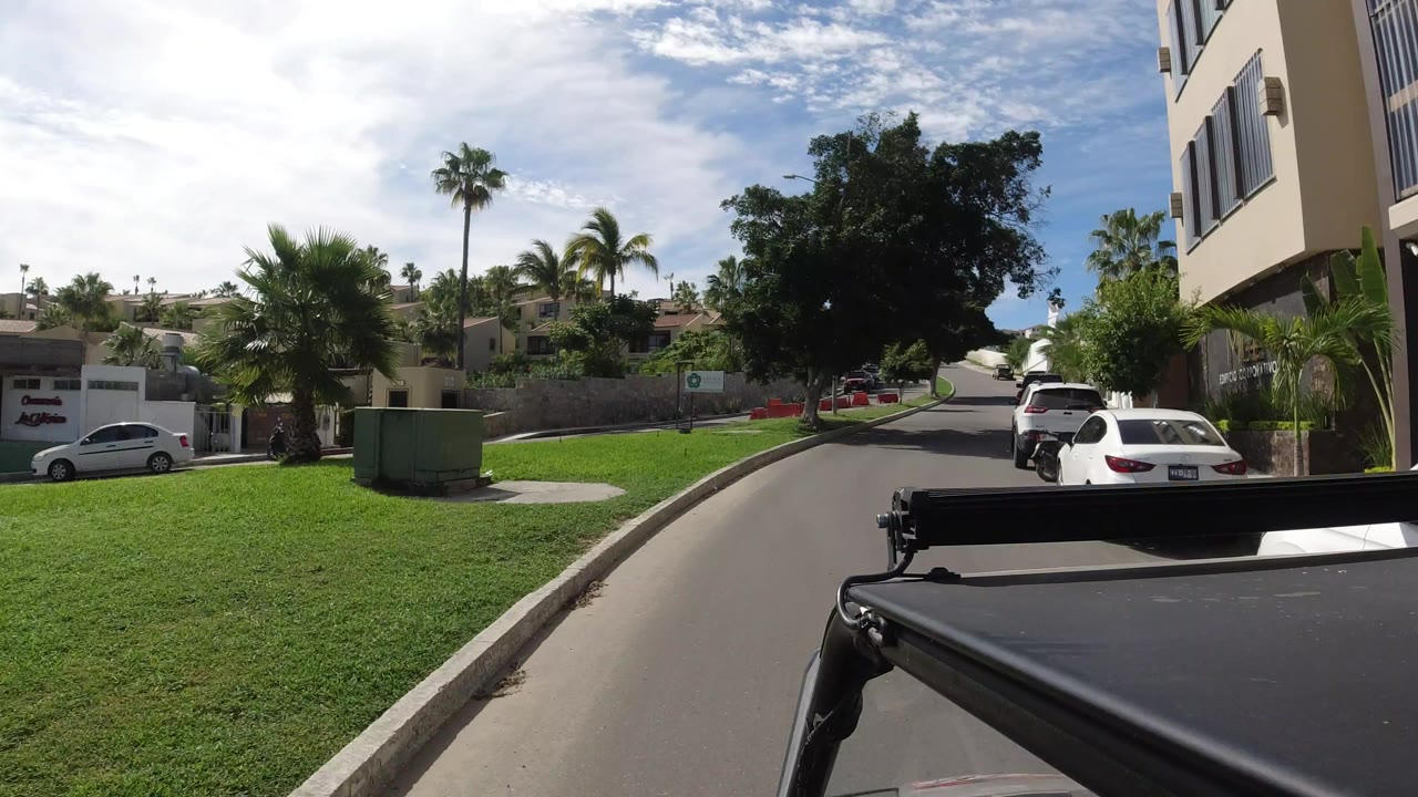 Vehicle-level view in Los Cabos, Mexico, with the camera moving along a quiet road past lawns, palms, a white car, modern homes, and no visible people.