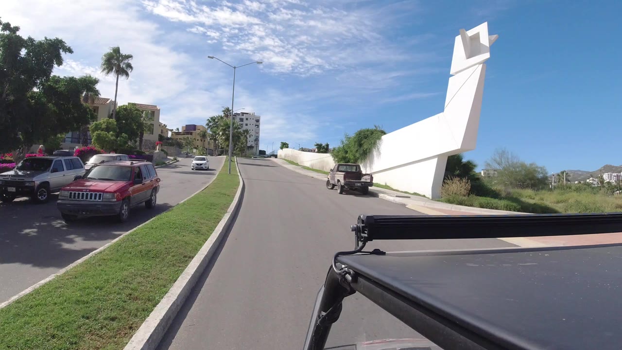 Driver's view on a sunny road with parked cars, palms, and a white wall.