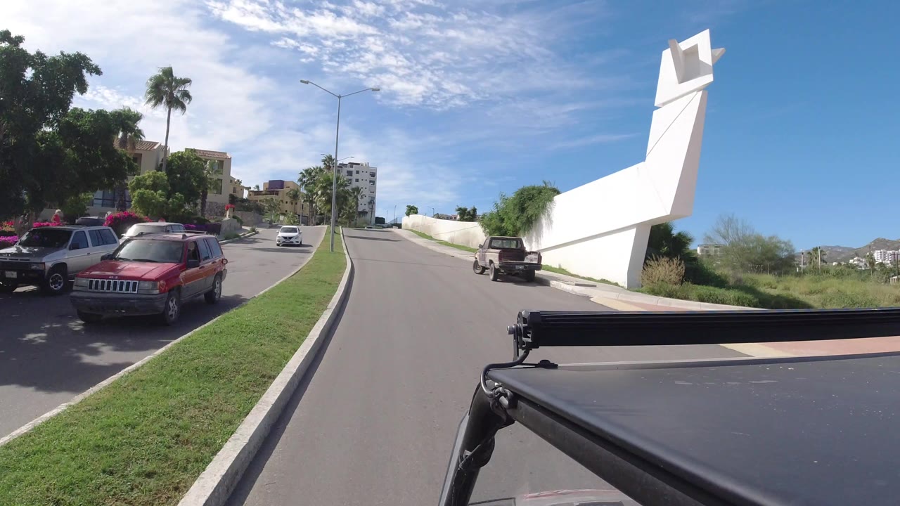 Los Cabos road view with a red parked SUV at left, a white perimeter wall at right, palm trees, distant buildings, and no visible people.