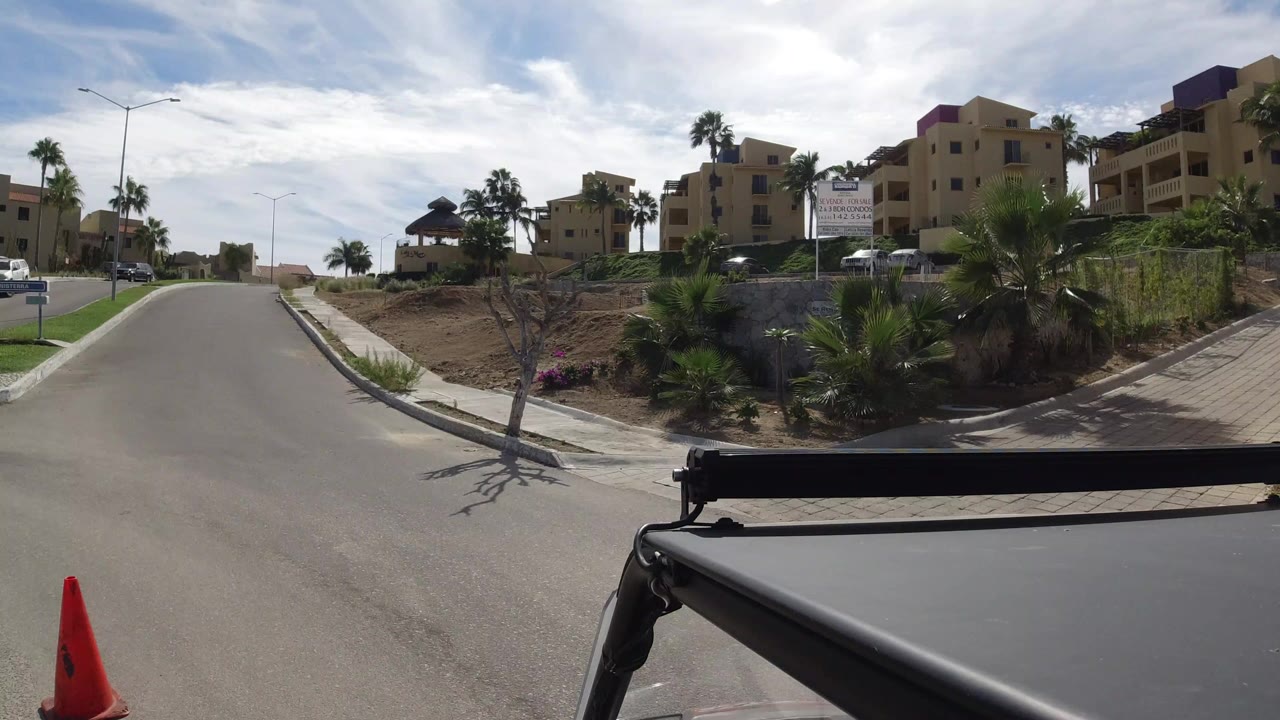 Street-level vehicle view in Los Cabos, Mexico, looking along a paved road with tan homes, palms, parked cars, and no visible people.