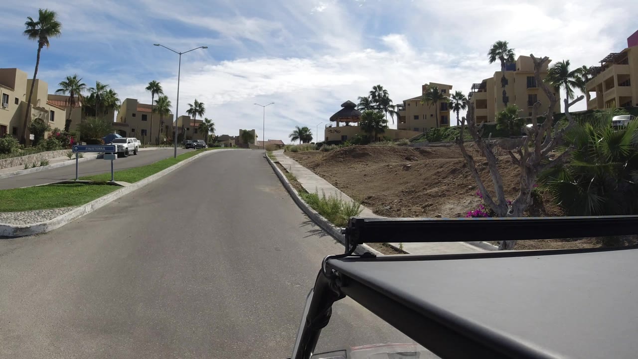 Curving residential road with palms, parked vehicles, and tan buildings.