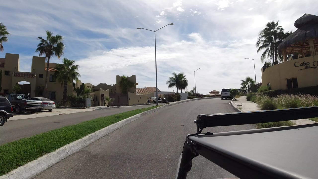 Street-level view in Los Cabos, Mexico, showing a curving residential road, palm trees, stucco buildings, a streetlight, parked vehicles, and no visible people.