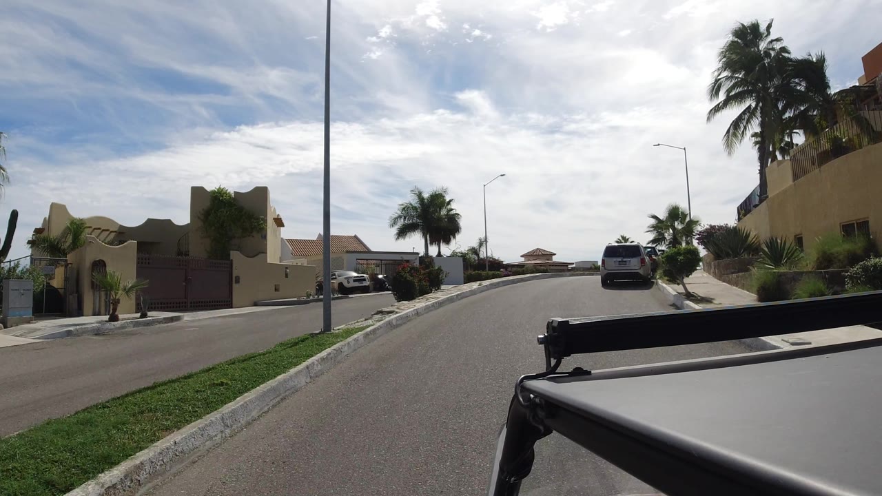 Residential street passing a tan building, palms, shrubs, and a parked white vehicle.