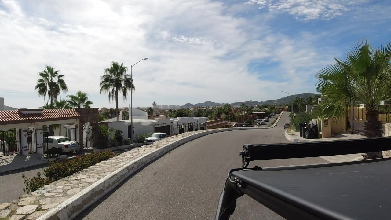 Opening view of a Los Cabos residential road with white homes, palm trees, parked cars, a stone curb, distant hills, and no visible people.