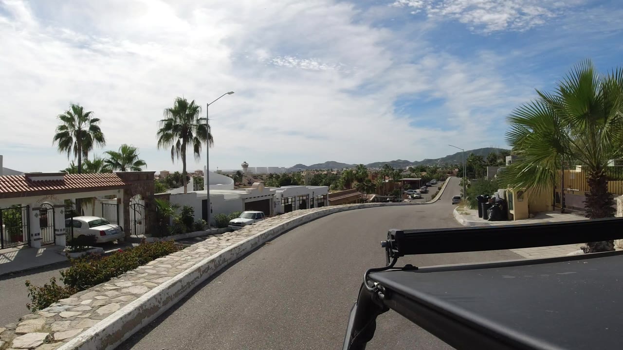 Los Cabos street view with a low white wall, gated homes, palm trees, parked vehicles, distant hills, and no visible people.