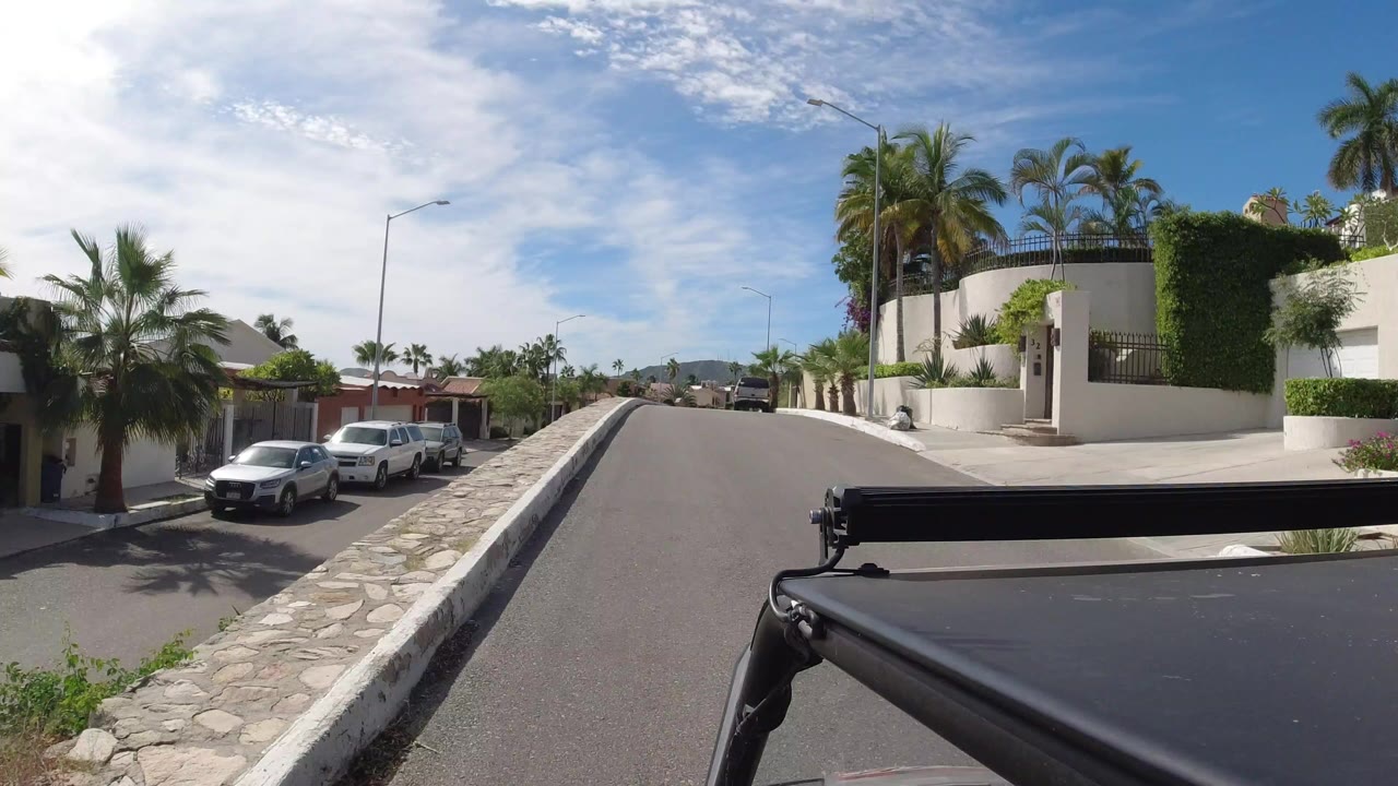 Sunny uphill residential road with parked cars and palm trees.