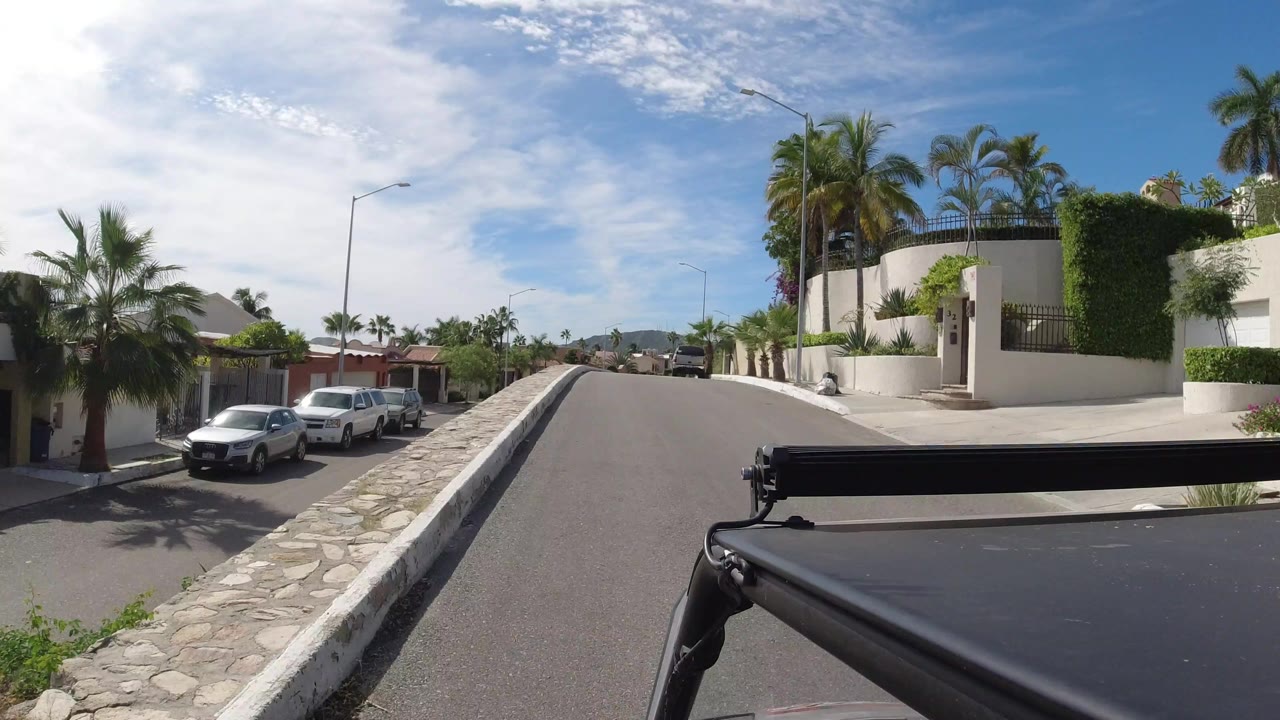 Los Cabos road view from a moving vehicle, with white residential walls, parked cars, palm landscaping, distant hills, bright sky, and no visible people.