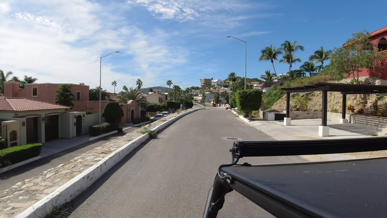 Sunny residential road with villas, palms, and vehicle foreground.