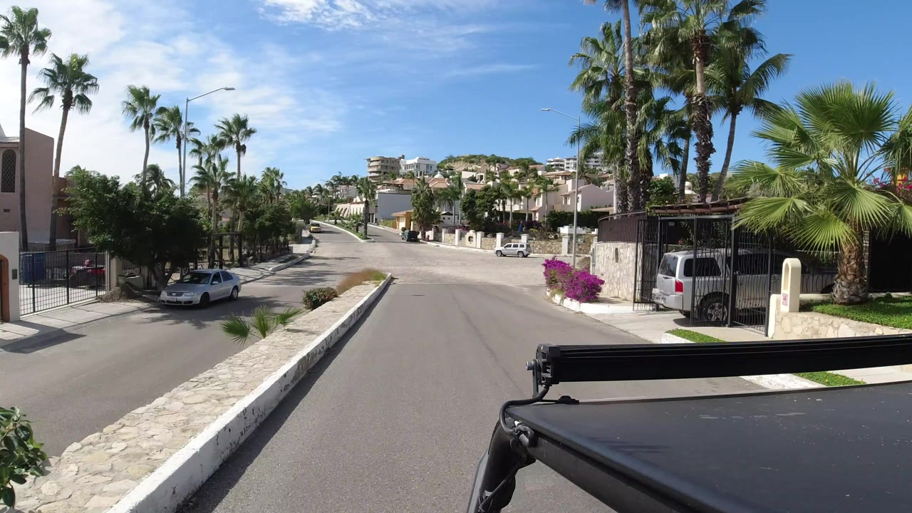 A Los Cabos residential road appears under bright sun with palm trees, walled homes, a white parked car, blue sky, and a black vehicle edge at the bottom of frame, with no visible people.