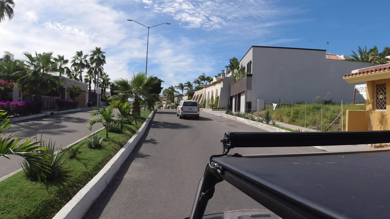 Opening driver-view frame in Los Cabos, Mexico, looking down a quiet street with palms, white homes, a van ahead, and no visible people.