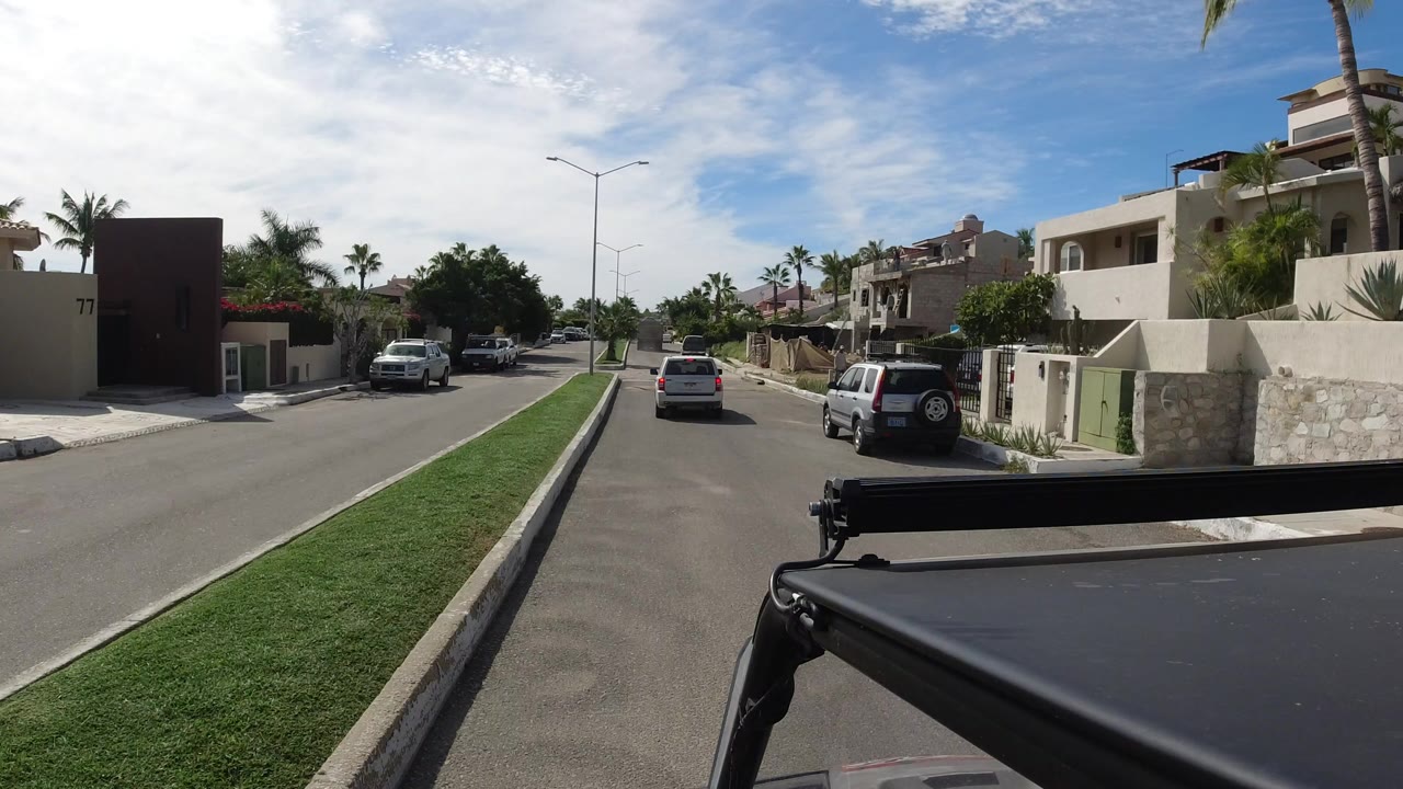 Vehicle-mounted view down a sunny residential street with homes and parked cars.