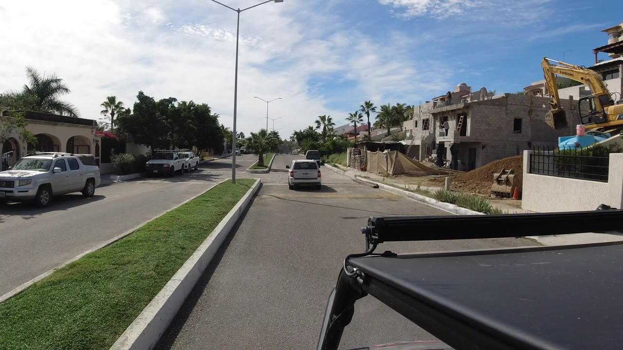 Los Cabos street view with the camera moving closer to the unfinished house, white pickups parked along the curb, a car in the lane, palms in the distance, and no clear faces visible.