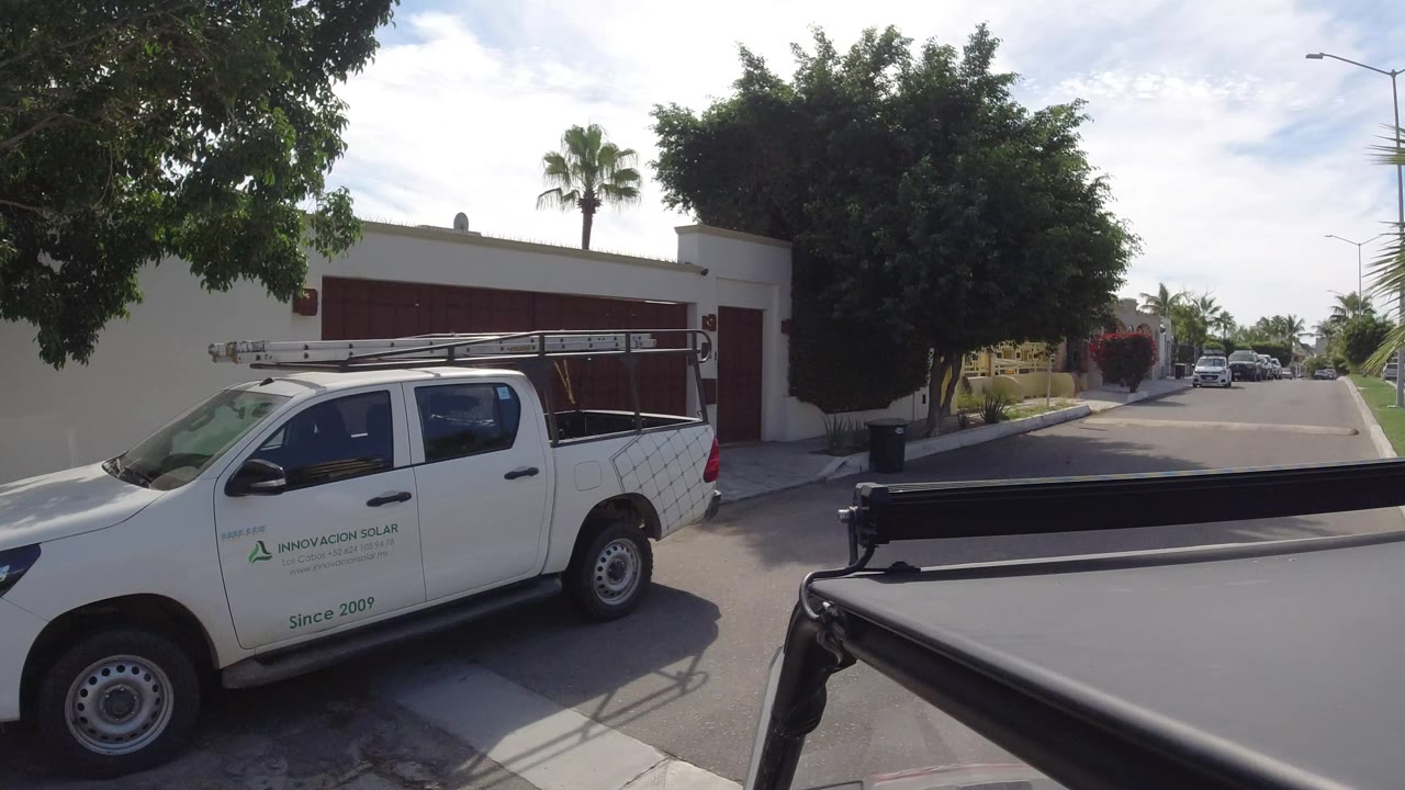 A white service pickup fills the left foreground as a white Los Cabos home wall, red garage gate, shade tree, palm, and bright street remain visible with no visible people.