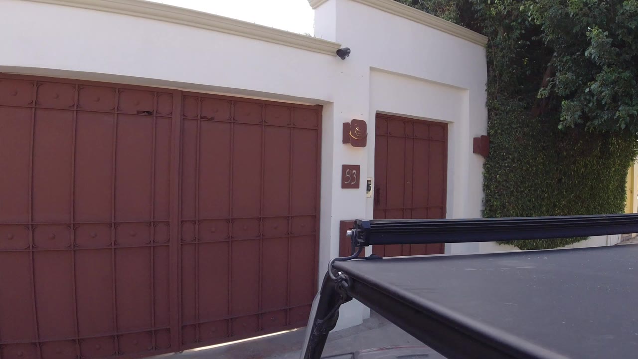 Opening frame of a Los Cabos house exterior with a red metal garage gate, white facade, hedge, and black vehicle roof in the foreground, with no visible people.