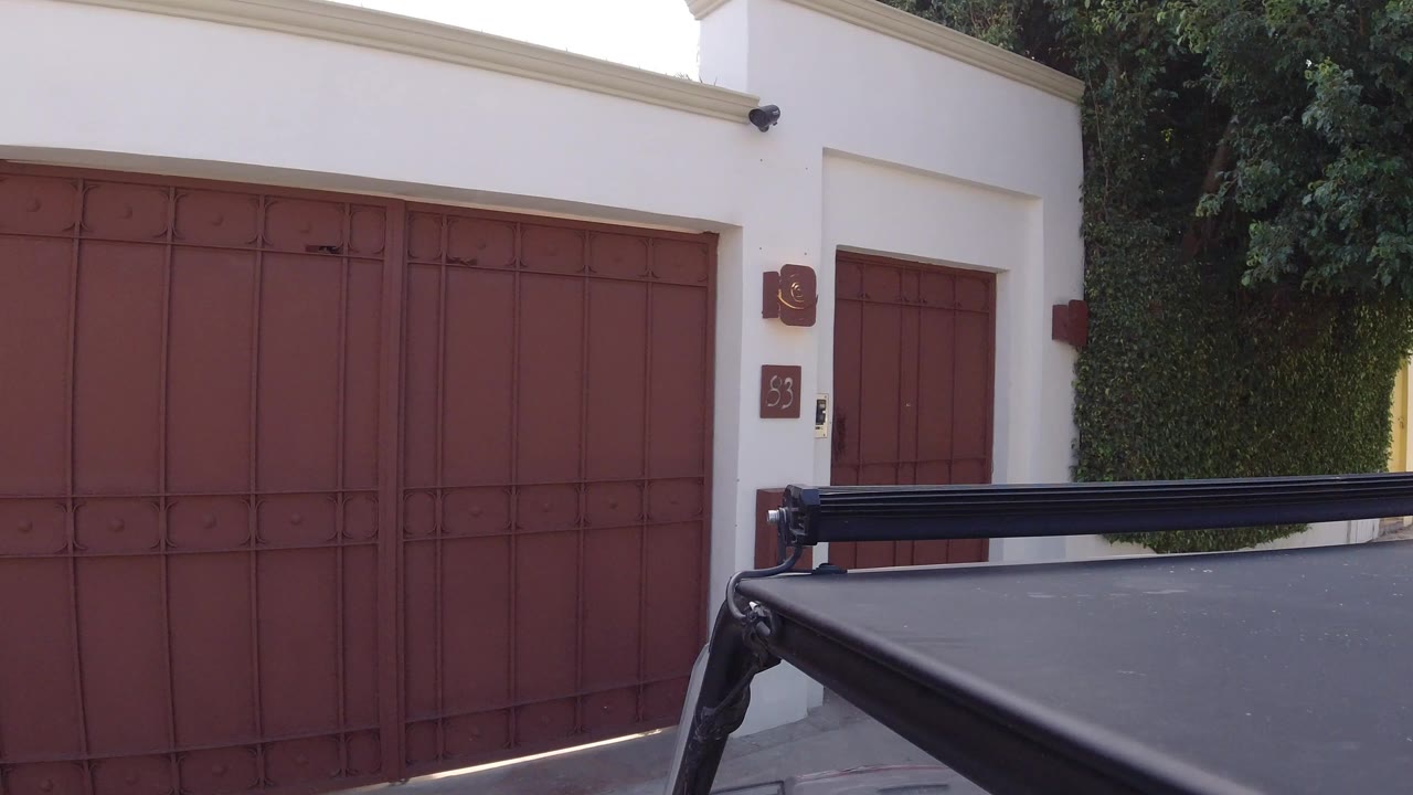Second frame shows the black vehicle roof and rack slightly farther across the lower frame, with the Los Cabos red garage gate and white wall still centered, and no visible people.