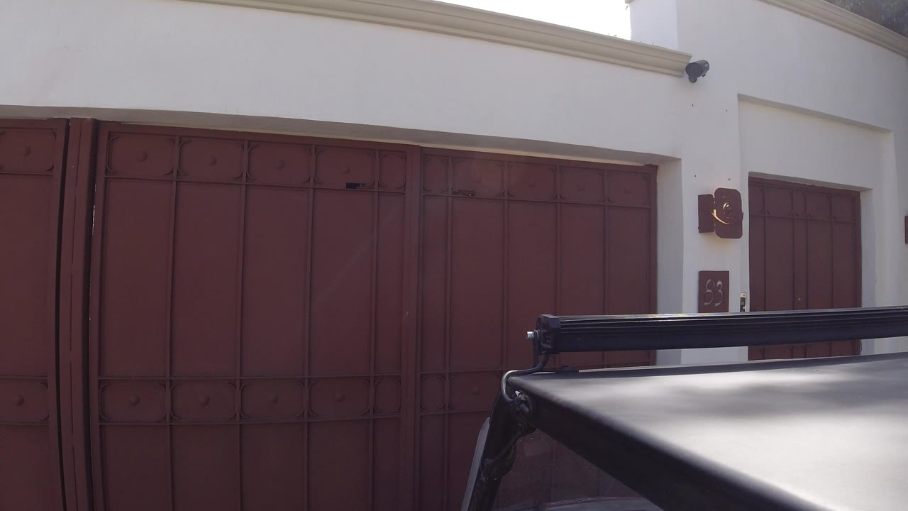 At 0:01 in Los Cabos, Mexico, the parked black vehicle remains in the lower foreground against broad red doors and a bright white wall, with no visible people.