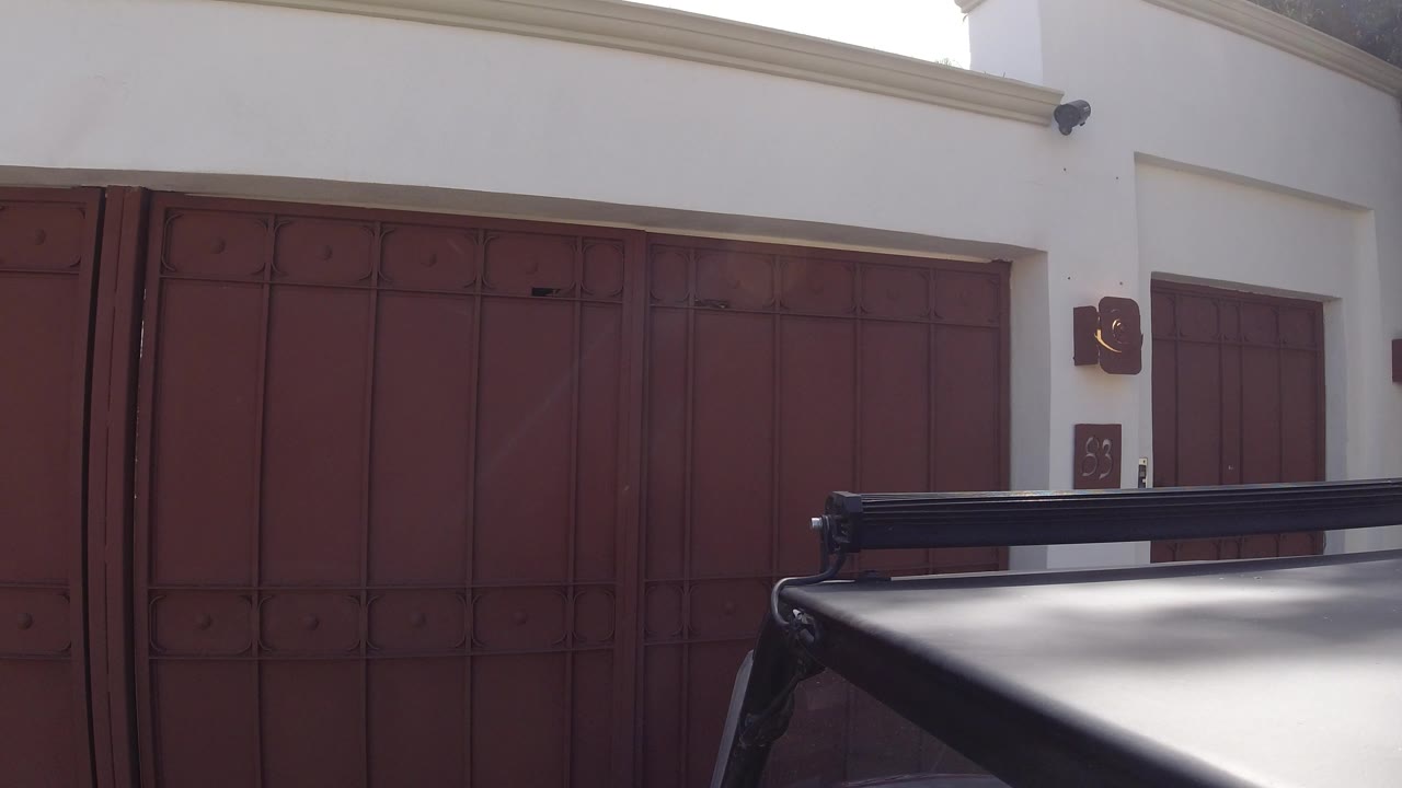Early frame in Los Cabos, Mexico with the red garage gate centered behind the dark car roof, white wall, and small side door, with no visible people.