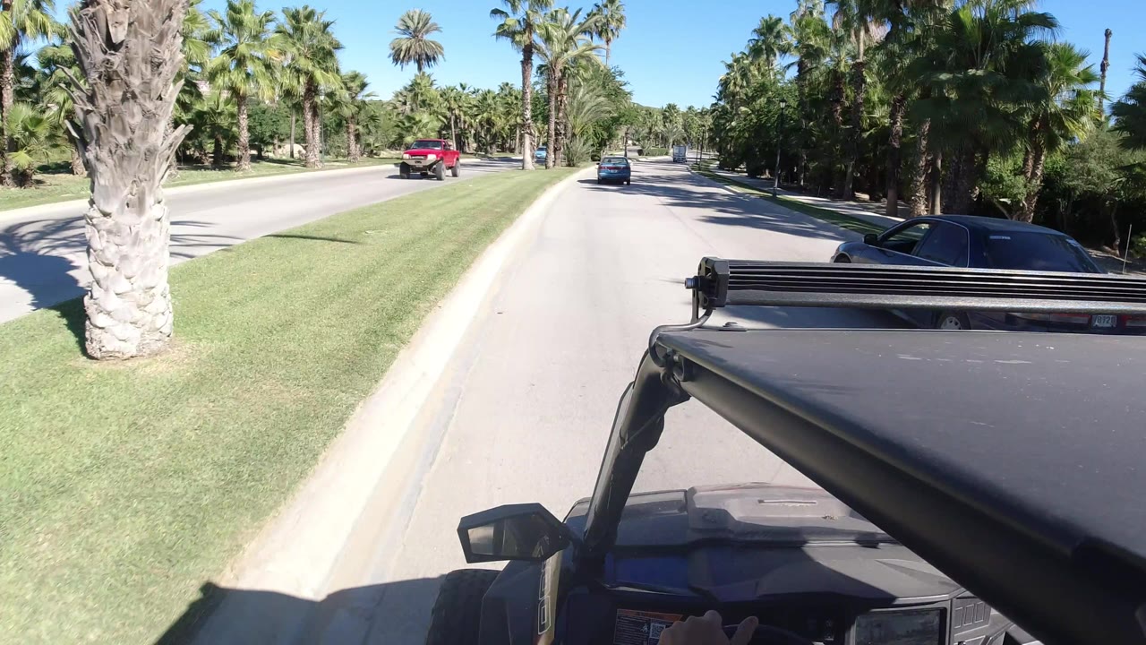 Forward-facing jeep view on a Los Cabos road as a red roadside object appears near the palm-lined curb, with cars in the distance and no visible people.