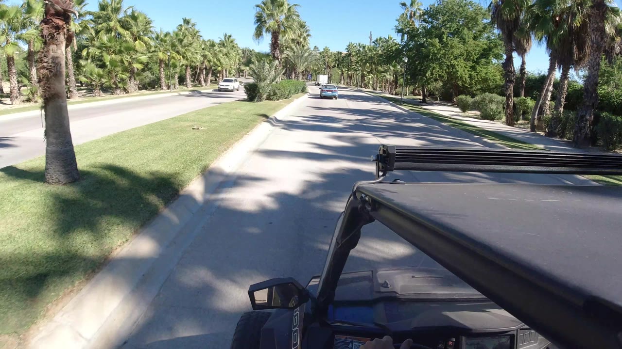 One second in, the Los Cabos road remains clear ahead, with the vehicle roof and side mirror in the foreground, palms on both sides, and no visible people.