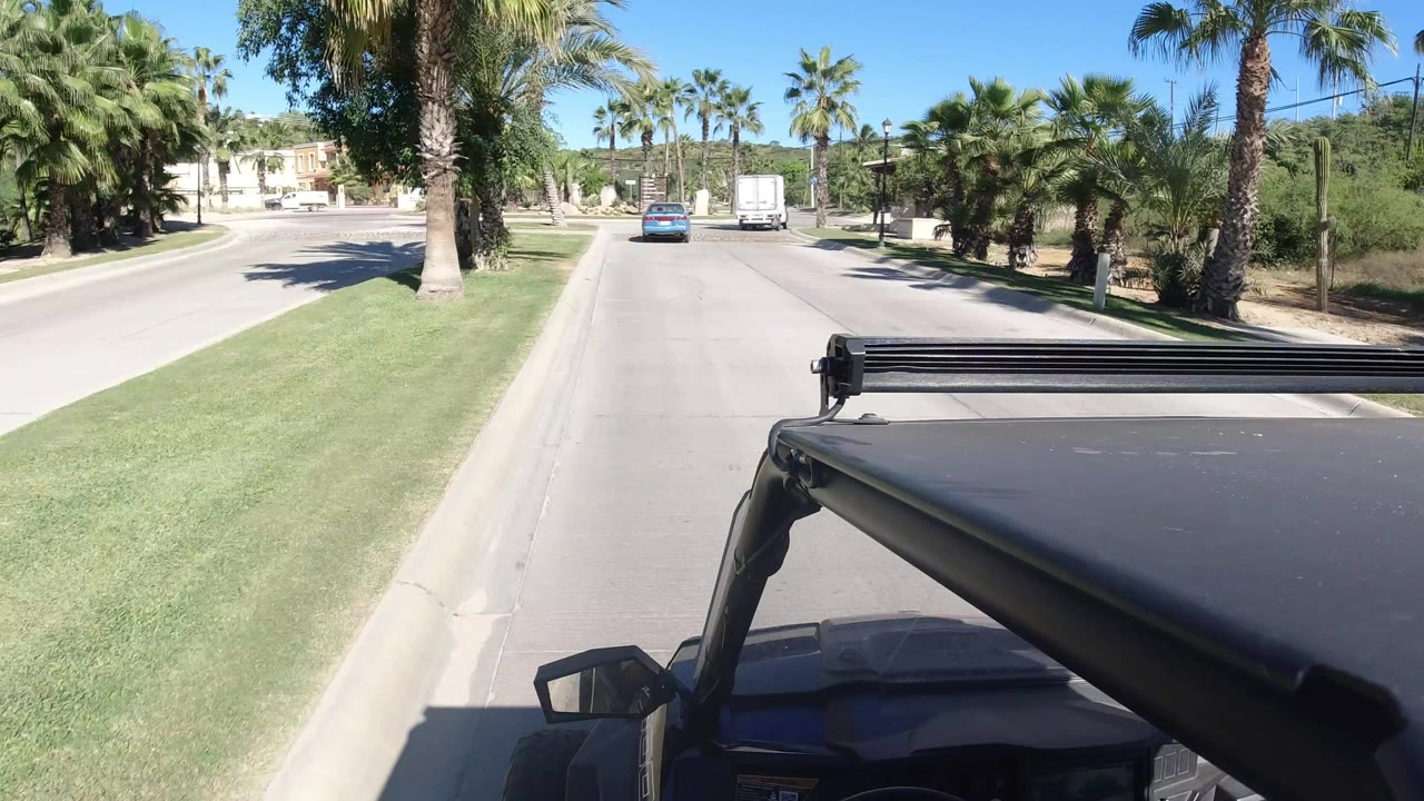 Forward view from a vehicle on a sunny palm-lined road behind a blue car and white truck.