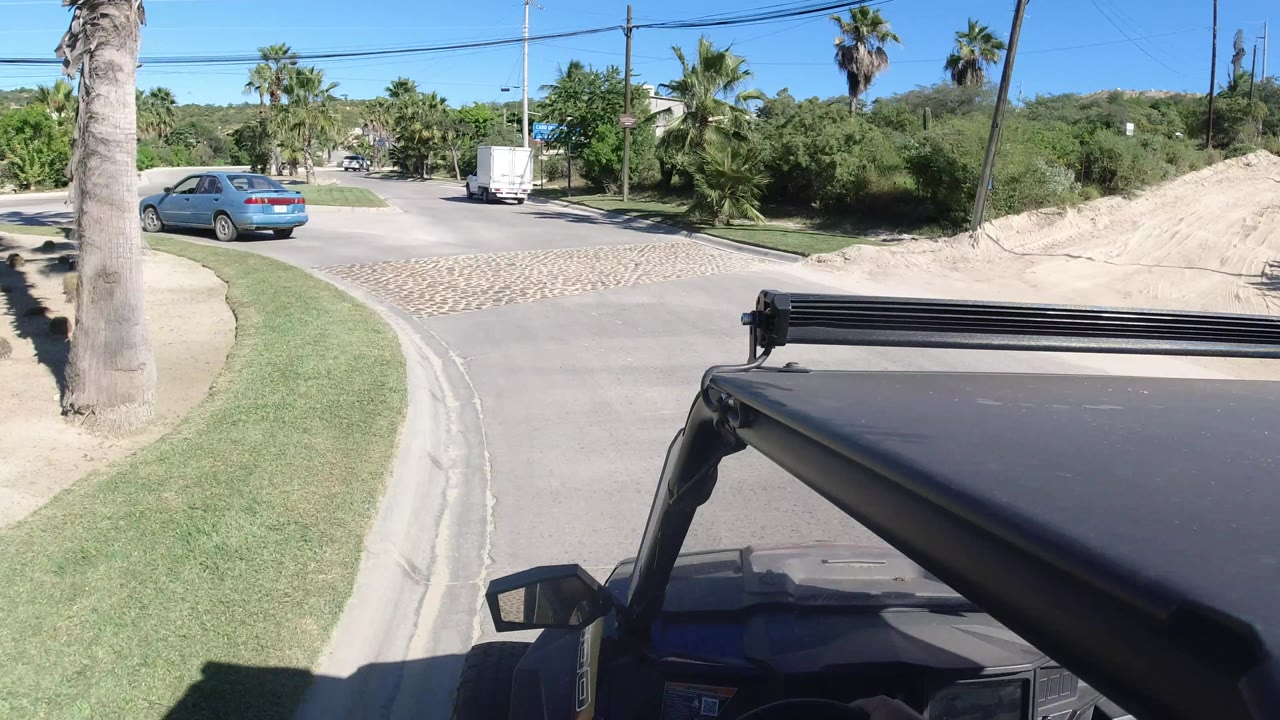 The vehicle continues behind a white truck on a Los Cabos street, with palms, low greenery, a road curve, overhead lines, and no visible people.