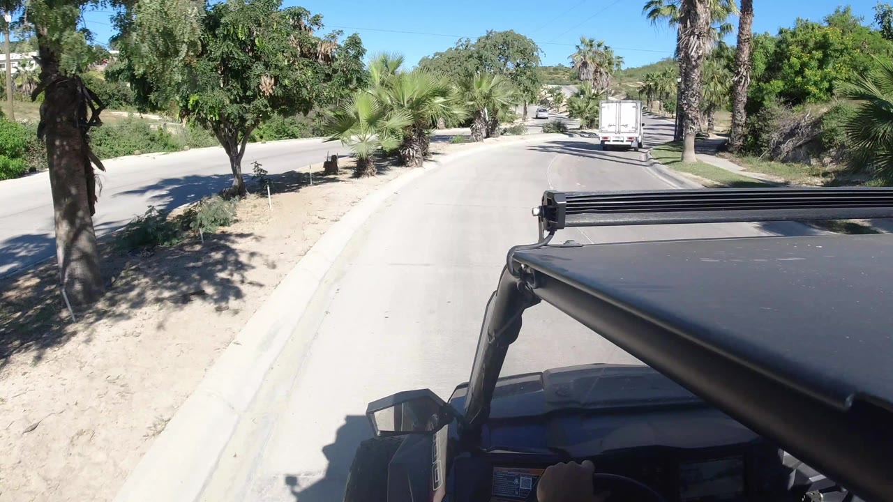 Vehicle-mounted view in Los Cabos, Mexico, showing the road continuing past palms and shrubs, with a white truck ahead and the filming vehicle's roof and bar filling the lower foreground.