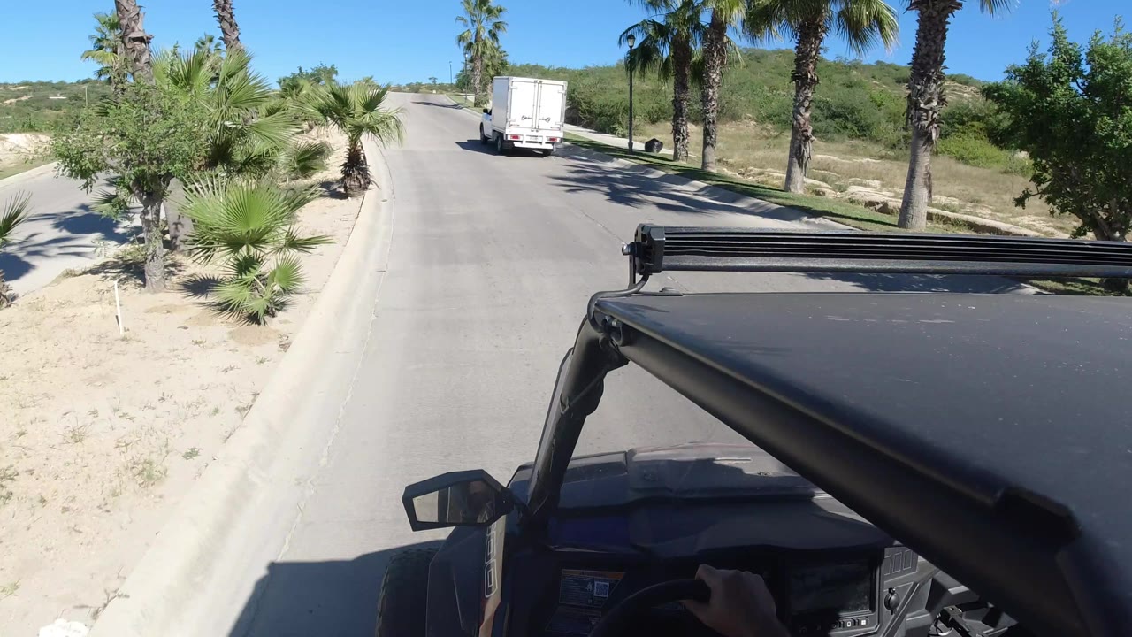 One second in, the white truck remains ahead of the black vehicle on the Los Cabos road, with palm trunks, low greenery, sandy shoulders, and no visible faces.