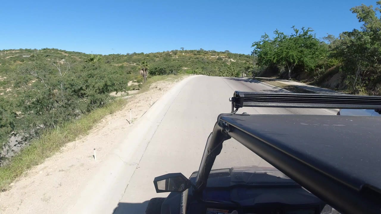 The Los Cabos roadway continues uphill between pale sandy shoulders and green brush, with the vehicle roof rack fixed across the lower right and no clearly visible people.