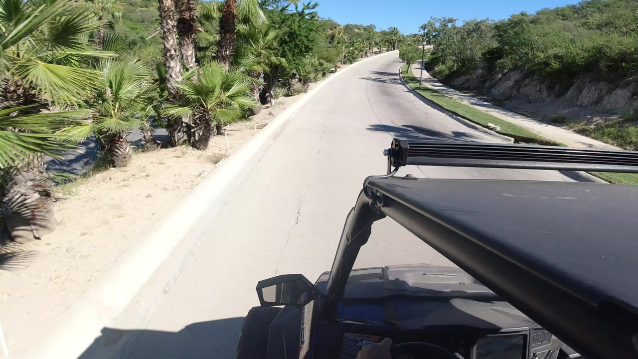 Black open-top vehicle foreground on a sunlit uphill road in Los Cabos, Mexico, with palms, shrubs, blue sky, and no visible faces.