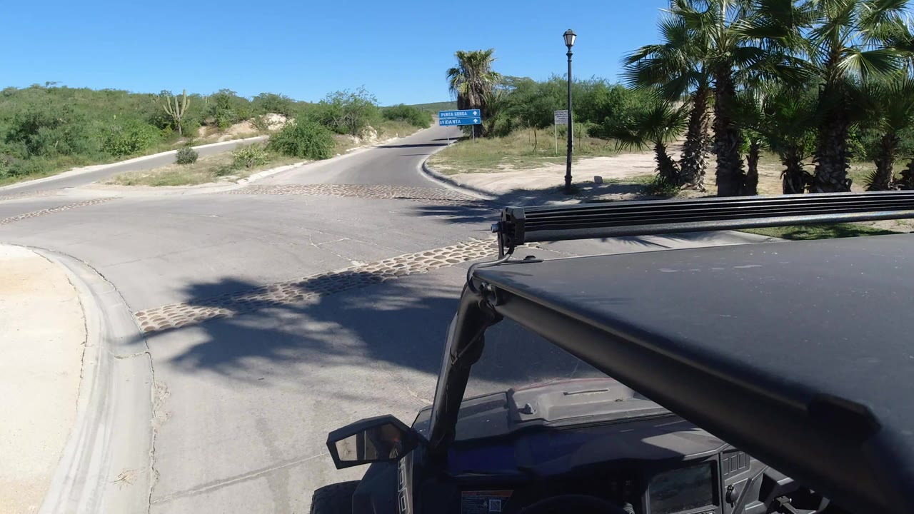 Vehicle-mounted view in Los Cabos, Mexico moving closer to the junction, with the black roof frame in the foreground and blue signs beside palms ahead.