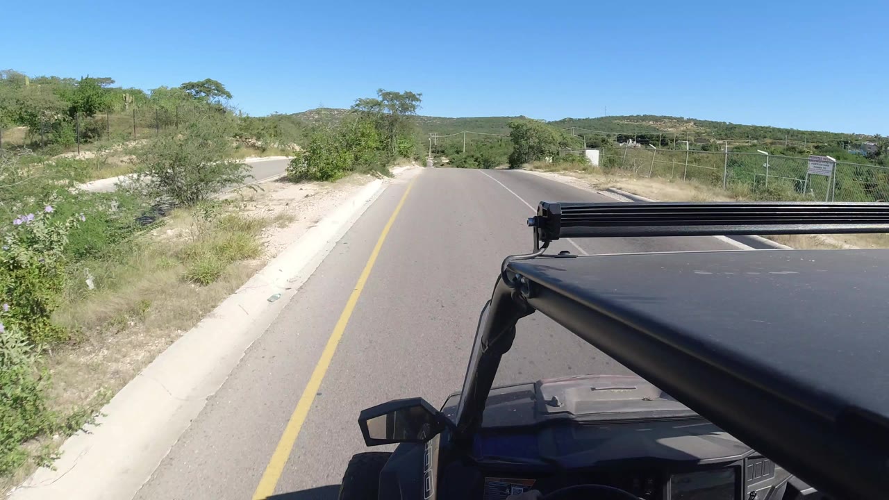 In Los Cabos, Mexico, the camera view stays over the Jeep roof as the road stretches ahead between a pale curb, dry shoulder, thick shrubs, and distant hills, with no visible people.