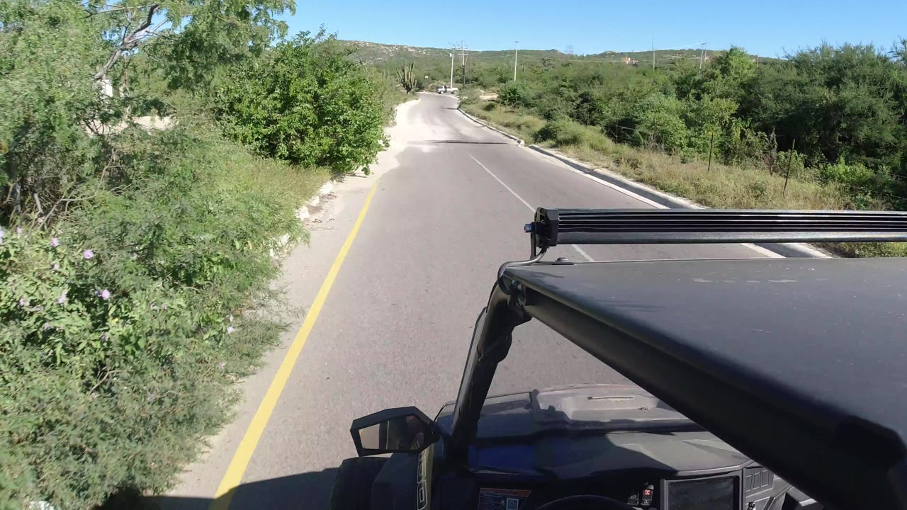 Forward-facing Los Cabos road view from above the black mini SUV hood, with the road stretching ahead between green vegetation, sandy edges, utility poles, distant hills, and no visible people.