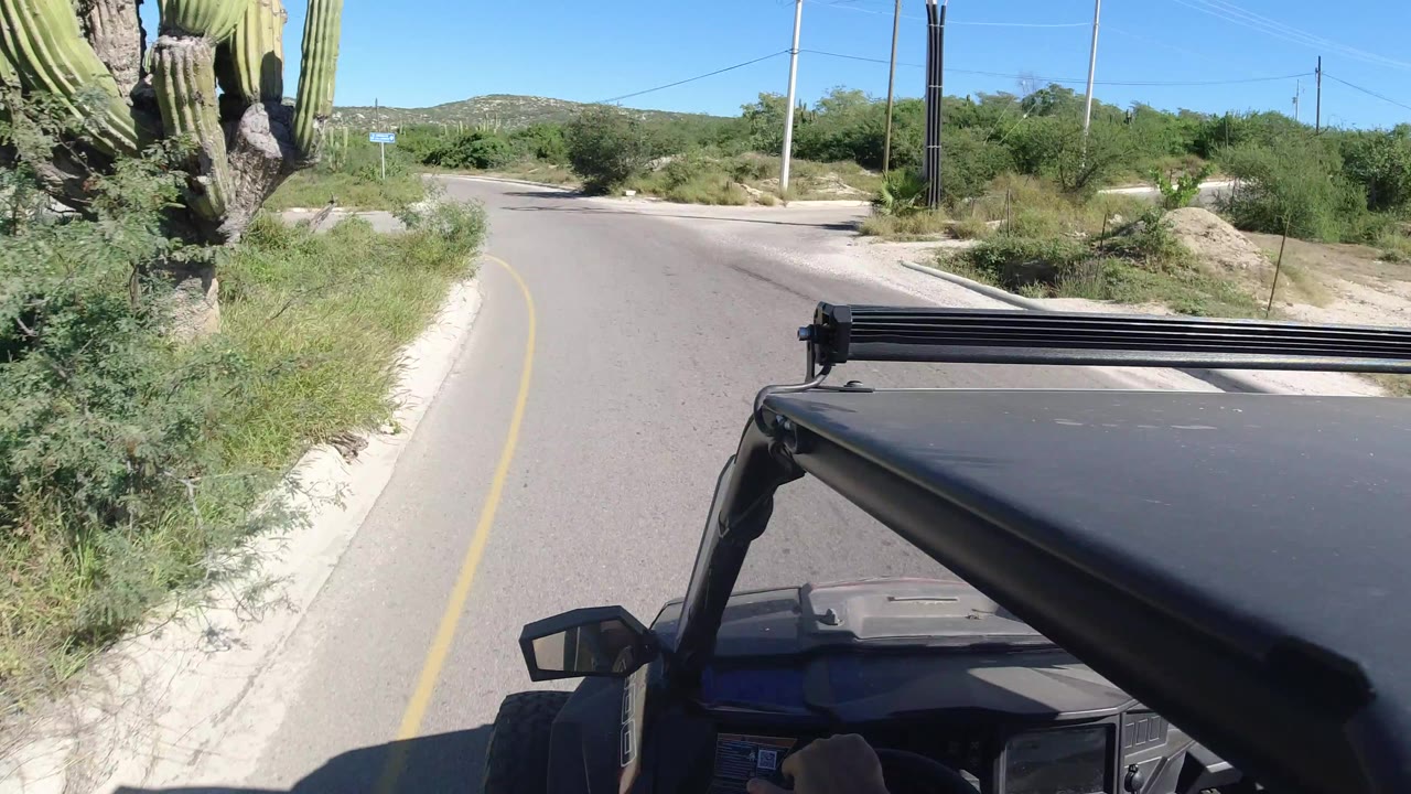 Vehicle-mounted view over the black roof as the paved road curves ahead in Los Cabos, Mexico, bordered by desert shrubs, cacti, power poles, distant hills, and no visible people.
