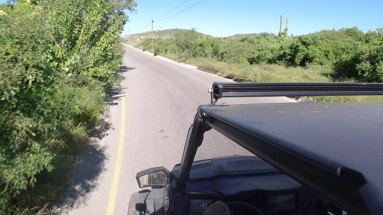 Forward vehicle-mounted view in Los Cabos with the black roof and light bar at the bottom, a straight paved road ahead, roadside brush, utility lines, and no visible face.