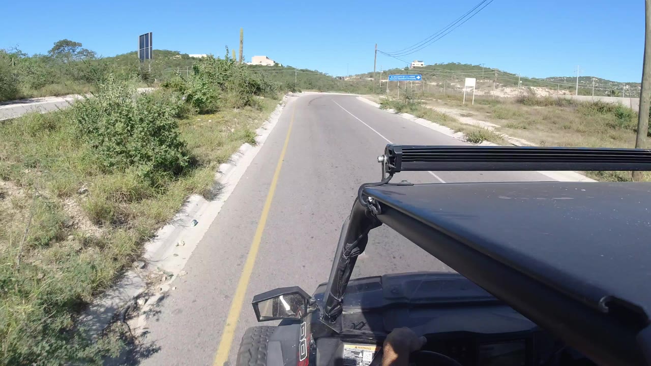Open vehicle driving on a paved desert road with scrub and low hills ahead.