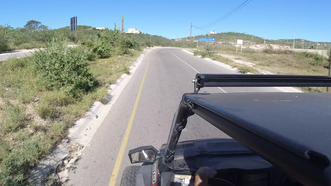 The vehicle continues along the paved Los Cabos road, with a brushy shoulder on the left, distant hillside buildings, utility lines, and no visible people.