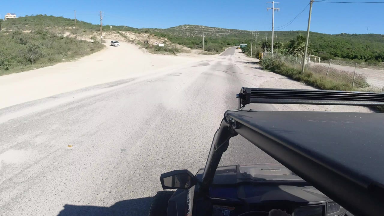 Mounted view from a black Jeep entering a pale dirt road in Los Cabos, Mexico, with sandy shoulders, scrub vegetation, utility poles, low hills, blue sky, and no visible people.