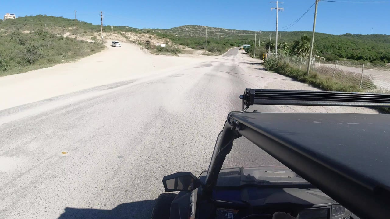Forward-mounted Jeep view in Los Cabos, Mexico, showing a sandy junction to the left, the main dirt road ahead, roadside brush, utility poles, and no visible people.